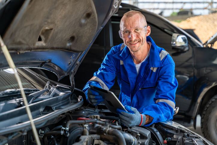 A smiling auto mechanic working on a car and holding a tab after completing auto mechanic certification