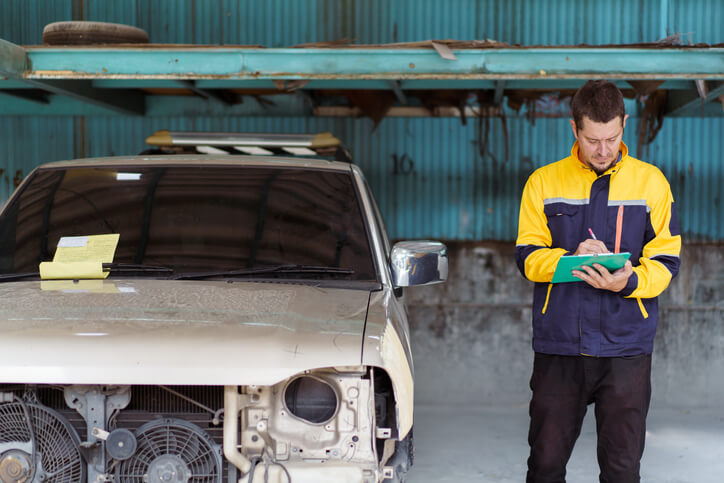 An auto body estimator in a workshop examining a beige car's condition with notes in hand for repair cost assessment