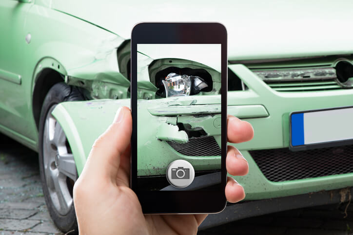 A close-up of a hand holding a smartphone, capturing the detailed damage on a green car's bumper for auto body estimating