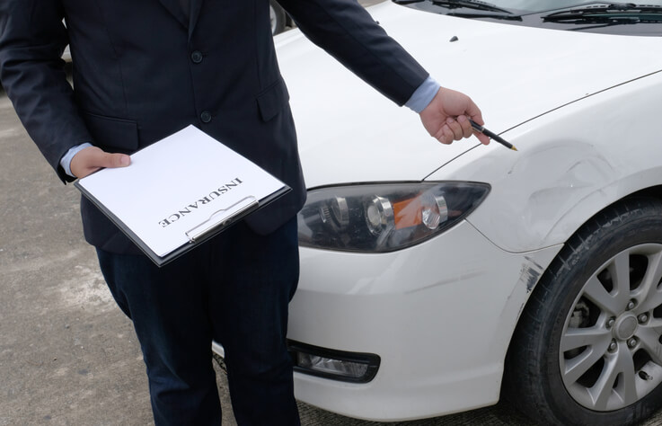 An auto body estimator in a suit inspecting damage on a white car's front bumper with a clipboard in hand