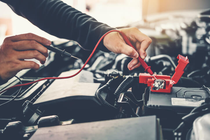 A male diagnostician examining a vehicle’s engine with a multimeter after graduating from auto mechanic school