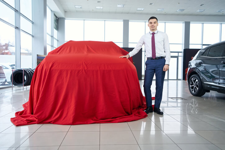 A young man is in a dealership after automotive school next to a car covered in red cloth.