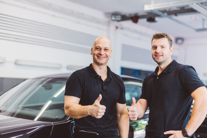 Two auto detailing training grads giving thumbs up in front of shiny cars