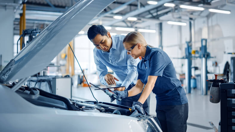 An auto mechanic school instructor guiding a student during hands-on training in a clean, well-organized shop.