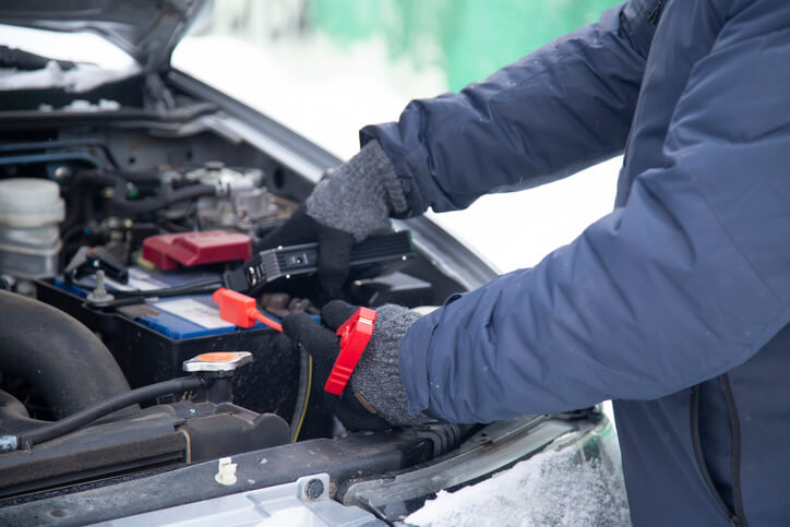 A student at an auto mechanic school performing a cold-weather battery test on a parked car in snowy conditions.