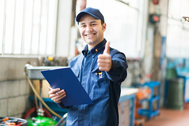 A graduate of an auto mechanic course in a Surrey training shop giving a thumbs up while holding a checklist clipboard.