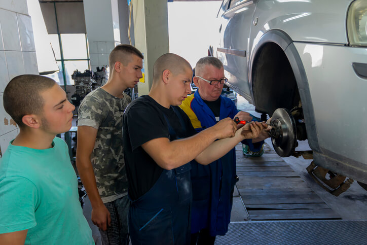 Auto mechanic instructor guiding a student on proper repair technique in a training garage.