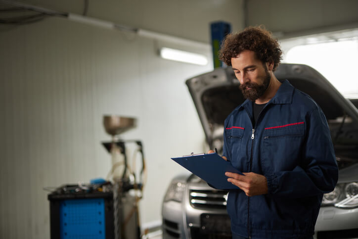 An automotive service technician program student reviewing a repair checklist on a clipboard during vehicle maintenance to ensure accuracy and completeness
