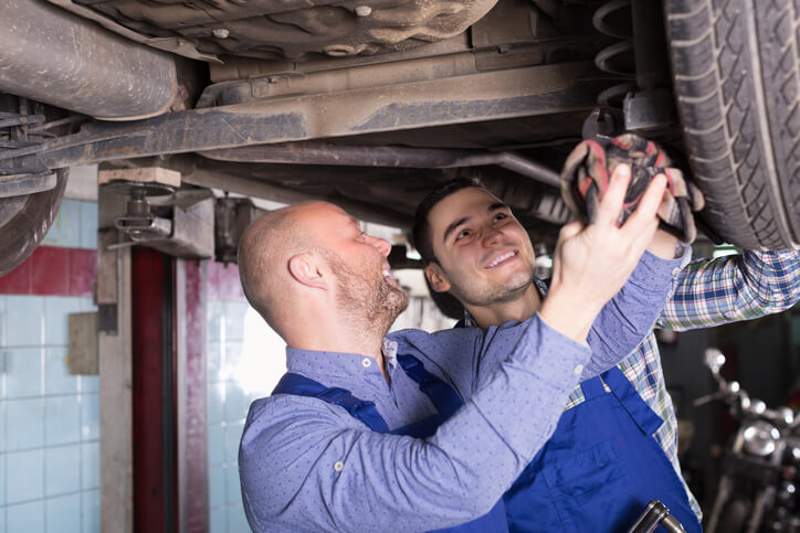 A senior and junior auto mechanics working together under a vehicle, demonstrating skill-level progression in an auto repair shop.