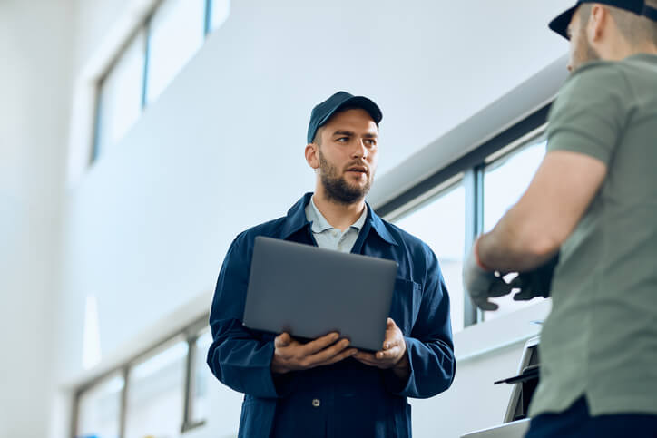 Two automotive technicians discuss diagnostics using a laptop before beginning repairs.