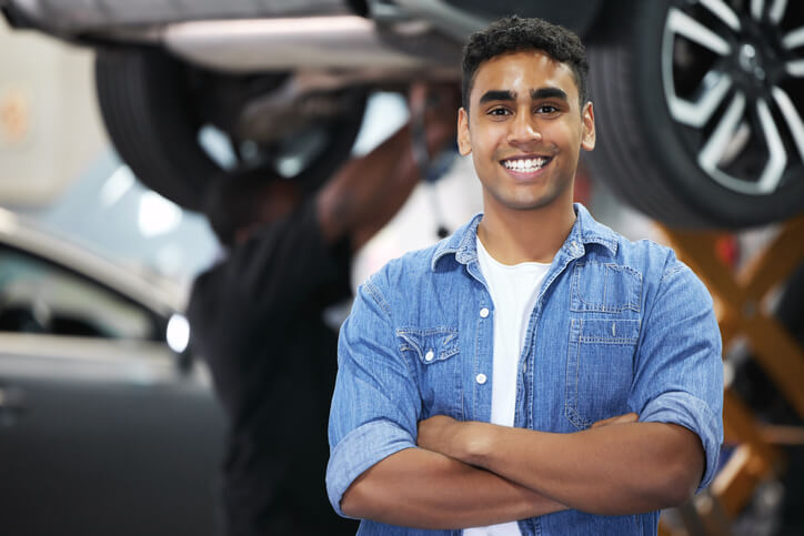 A young mechanic standing in an auto workshop with arms folded, wearing a work uniform.