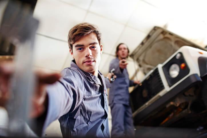 An automotive student assisting a mechanic checking a timing chain and guides for wear