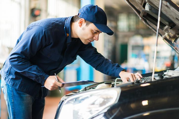 Automotive technician inspecting a vehicle under the hood while holding a clipboard